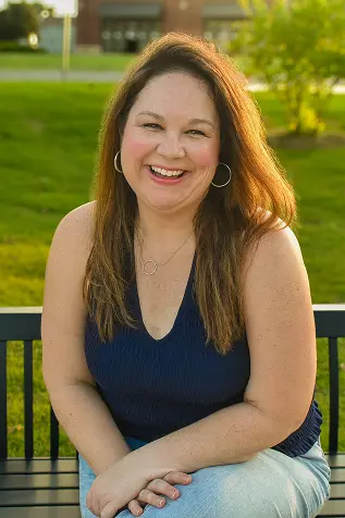 Melissa Talkington, a woman sitting and smiling on a park bench wearing a navy blue tank and blue jeans. She has brown hair and brown eyes. Her legs are crossed and her hands are resting on her knees. The sun is shining.
